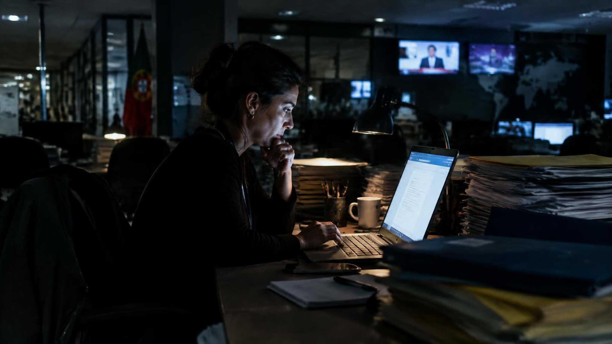 Journalist writing notes surrounded by folders in a dim newsroom